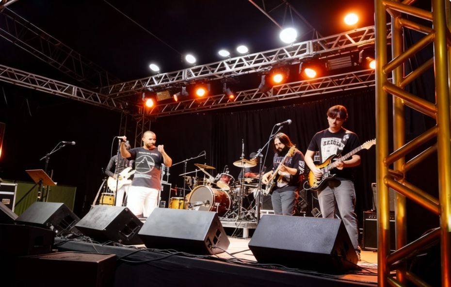 Fotografia de uma banda de rock composta por quatro integrantes tocando em um palco iluminado por refletores laranjas e brancos durante a noite.