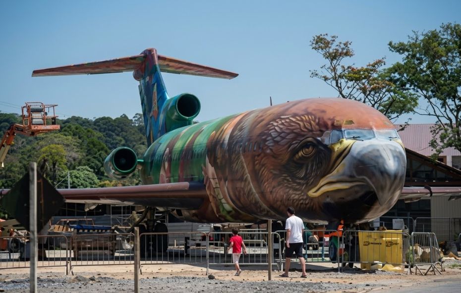 Fotografia em ângulo frontal e de baixo para cima de um avião Boeing 727 pintado com a face de um gavião em arte de grafite. Duas pessoas caminham na frente da aeronave, que está em uma praça cercada por grades metálicas e árvores ao fundo.