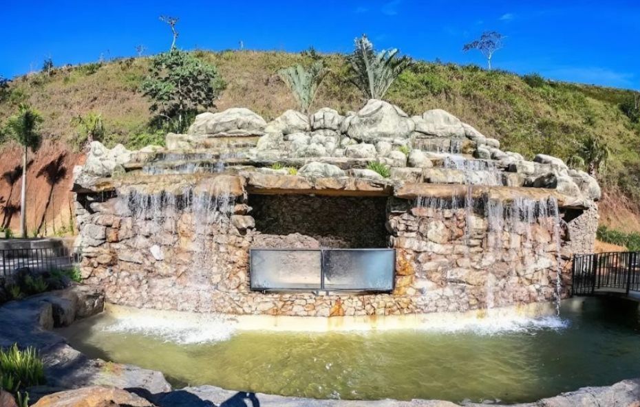 Fotografia de uma cascata artificial construída com pedras rústicas, apresentando múltiplas quedas d'água que deságuam em uma piscina rasa sob um céu azul limpo.