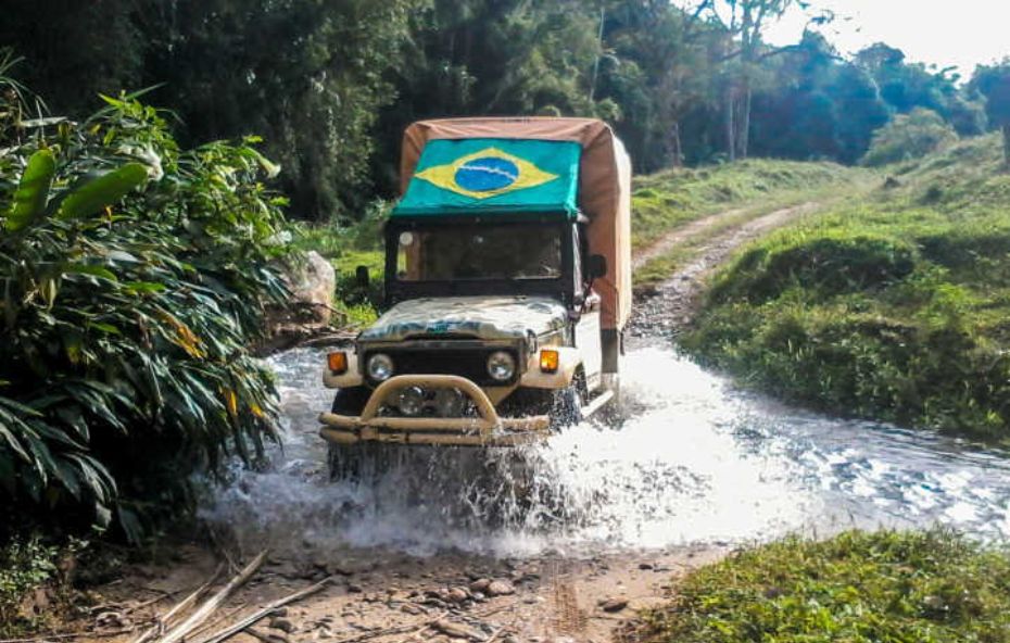 Fotografia de um veículo off-road bege com uma bandeira do Brasil na cobertura, atravessando um riacho em uma estrada de terra cercada por vegetação densa e verde.