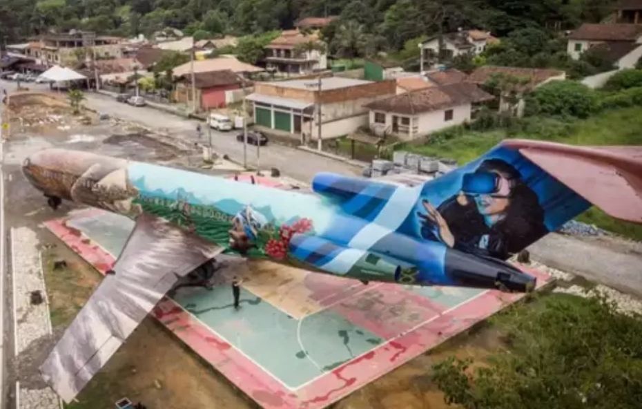 Vista aérea de um avião comercial de grande porte posicionado em uma praça urbana, com a fuselagem inteiramente decorada por grafites coloridos que incluem paisagens naturais e uma pessoa utilizando óculos de realidade virtual na cauda.