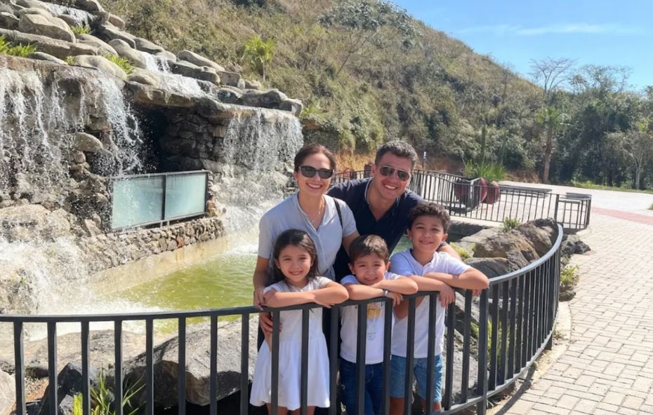 Uma família composta por pai mãe e três filhos sorri para a foto em um mirante com grades pretas tendo ao fundo a queda d'água da Cascata de Ipiabas com suas rochas decorativas e águas cristalinas sob a luz do sol.