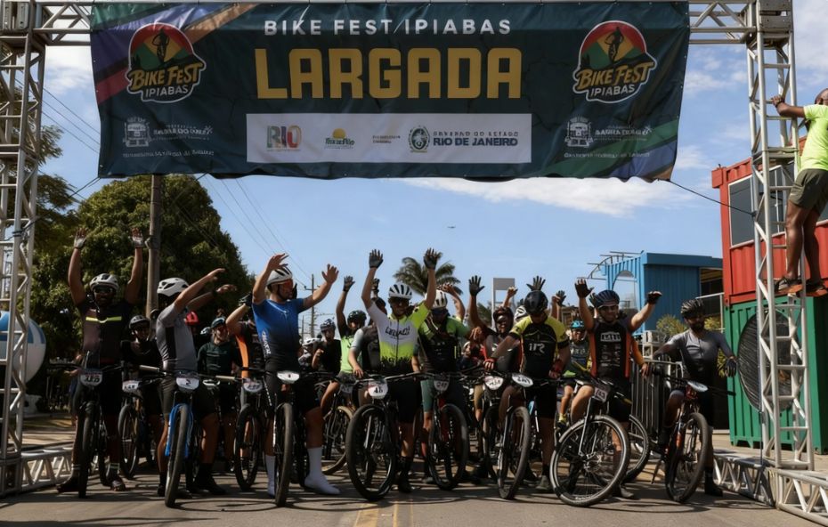 Fotografia de um grupo de ciclistas com capacetes e roupas esportivas posicionados sob uma faixa azul com a inscrição Largada Bike Fest Ipiabas, com os braços erguidos em sinal de celebração.