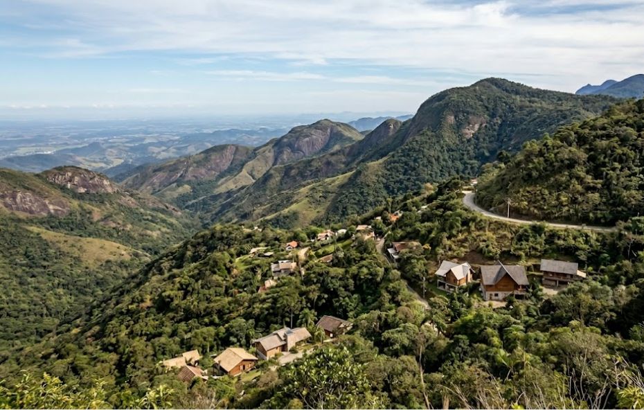 Uma vista aérea abrangente revela uma pequena vila aninhada nas encostas verdejantes da Serra do Mar, com picos íngremes e um vale vasto sob um céu parcialmente nublado no Rio de Janeiro.