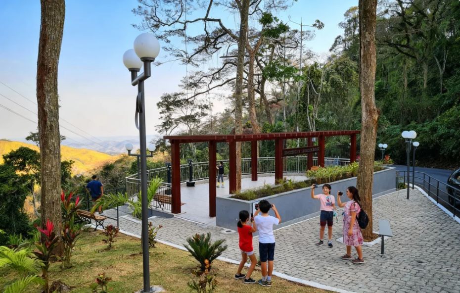 Fotografia de um mirante estruturado com pergolado de madeira e piso de pedra, onde pessoas fotografam um vale coberto por Mata Atlântica sob o céu claro.