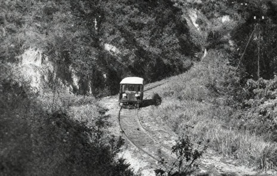 Fotografia em preto e branco de um pequeno veículo ferroviário motorizado conhecido como auto de linha trafegando sobre trilhos singulares. O trilho faz uma curva acentuada por entre encostas íngremes e vegetação densa em uma região de serra.