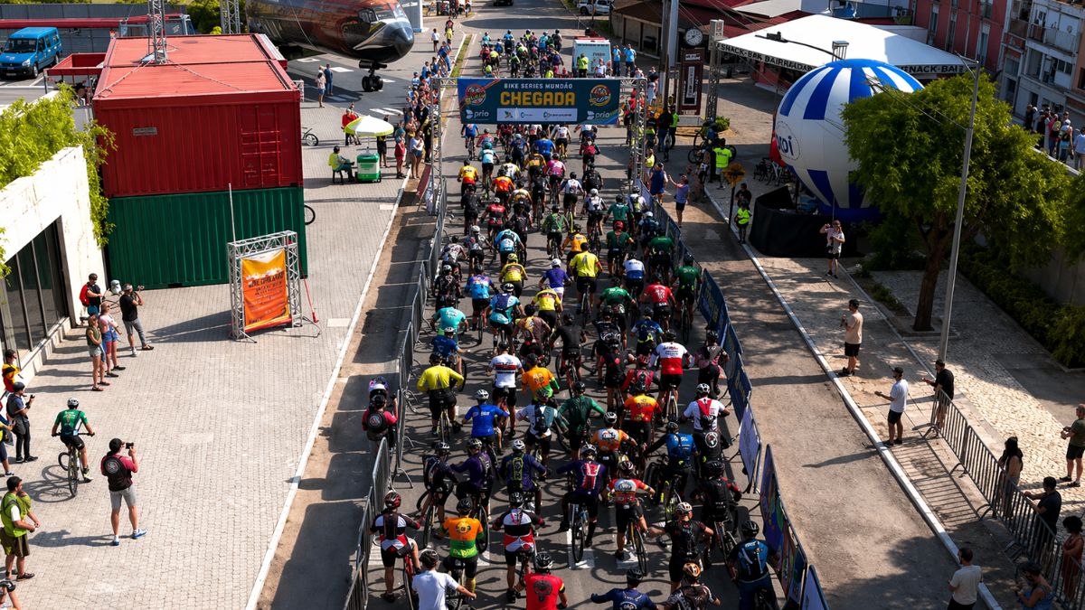Uma fotografia aérea de ângulo elevado capturando a linha de chegada de uma competição de ciclismo em Ipiabas. No centro da imagem uma multidão de ciclistas vestindo camisas coloridas e capacetes de proteção avança em direção a um pórtico metálico que exibe a palavra Chegada em letras amarelas sobre um fundo azul. O pórtico também contém logotipos de patrocinadores e o nome do evento Bike Series Mundão. A estrada é cercada por grades de metal cinza que separam os competidores do público e das áreas técnicas. À esquerda da pista há containers de carga nas cores vermelho e verde e algumas pessoas observando a prova. Ao fundo à esquerda a parte frontal de um avião Boeing está posicionada perto da via. À direita da pista vê-se um grande balão inflável listrado de azul e branco. O dia está ensolarado e a luz projeta sombras nítidas dos ciclistas no asfalto.