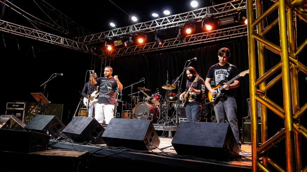 Fotografia de uma banda de rock blues se apresentando em um palco de estrutura metálica sob iluminação vibrante de refletores laranjas e brancos. O vocalista, ao centro, veste uma camiseta preta do Pink Floyd e bermuda branca e gesticula para o público. À direita, um baixista e um guitarrista concentrados tocam seus instrumentos, enquanto um baterista completa o conjunto ao fundo. Caixas de retorno pretas estão alinhadas na frente do palco e uma grade metálica amarela emoldura a lateral direita da imagem, criando uma atmosfera de show ao vivo em um ambiente noturno.
