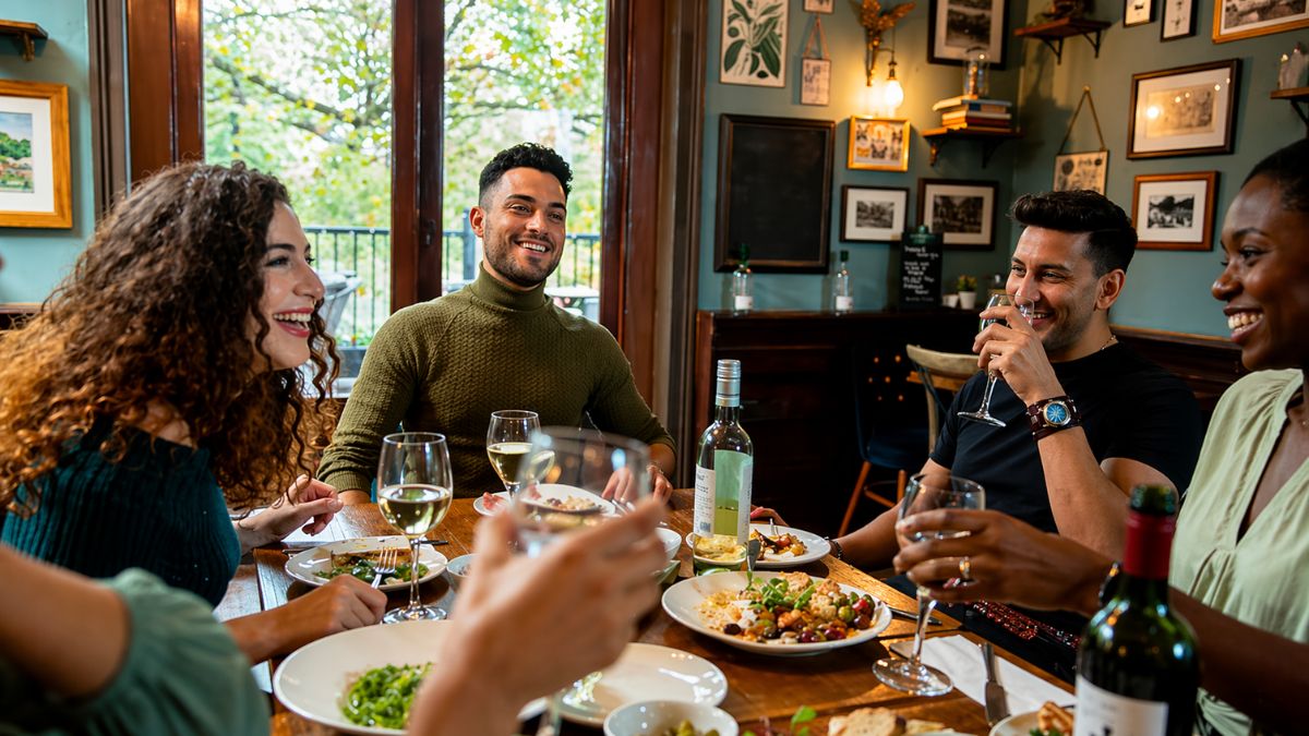 Fotografia de um grupo diversificado de quatro amigos sorrindo e conversando ao redor de uma mesa de madeira em um restaurante aconchegante. Eles desfrutam de uma refeição sofisticada com pratos coloridos, incluindo saladas e massas, acompanhados de taças de vinho branco e garrafas sobre a mesa. O ambiente possui paredes verdes decoradas com quadros e prateleiras de madeira, criando uma atmosfera de bistrô elegante. Grandes janelas ao fundo revelam árvores verdes, integrando a natureza ao espaço interno iluminado de forma suave.