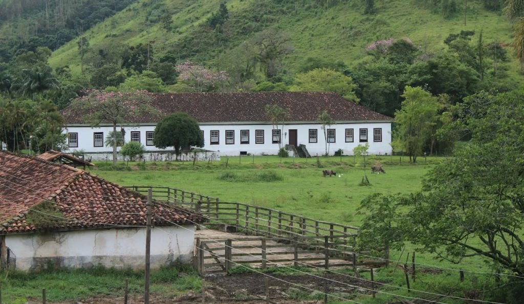 Fotografia de uma fazenda colonial com um casarão branco de janelas escuras e telhado de barro, situada em um vale verdejante com gado pastando à frente e montanhas ao fundo.