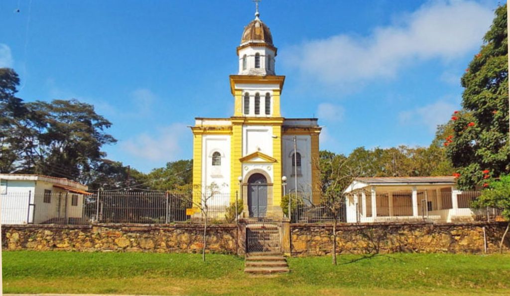 Fotografia frontal da Igreja de Nossa Senhora da Piedade, uma edificação histórica com torre central, paredes brancas e detalhes em amarelo, situada atrás de um muro de pedras sob céu azul com poucas nuvens.