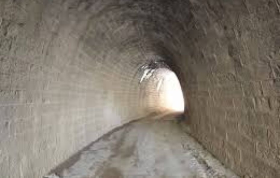 Fotografia do interior de um túnel ferroviário antigo construído com blocos de pedra e alvenaria aparente. A perspectiva mostra o teto em arco e as paredes laterais texturizadas que levam a uma saída iluminada pela luz do dia ao fundo criando um forte contraste entre a penumbra interna e o exterior.