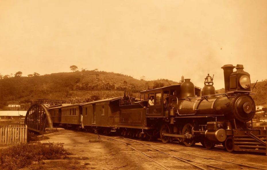 Fotografia antiga em tons de sépia mostrando uma locomotiva a vapor com o número sete na frente puxando vagões de passageiros e carga em uma ferrovia no Brasil. Ao fundo observa-se uma colina com vegetação rasteira e uma estrutura de ponte metálica à esquerda da composição ferroviária.