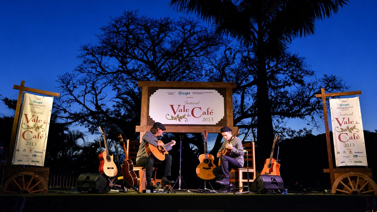 Fotografia noturna de um palco ao ar livre sob um céu azul profundo ao entardecer. Dois músicos estão sentados em cadeiras de madeira tocando violões de forma concentrada. Entre eles e nas laterais do palco, há outros violões posicionados em suportes. Ao centro do palco, um grande painel de madeira exibe o logotipo do Festival Vale do Café dois mil e treze. Dois banners verticais com a mesma identidade visual ladeiam a estrutura. Grandes árvores e palmeiras formam a silhueta escura ao fundo, criando uma atmosfera cultural e bucólica em meio à natureza.