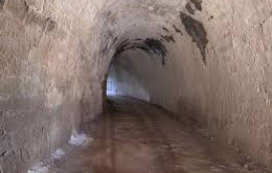 Fotografia do interior de um túnel ferroviário antigo com paredes e teto em arco feitos de pedra bruta e alvenaria de tijolos. O chão de terra batida reflete uma leve umidade e a passagem faz uma curva suave para a esquerda revelando uma luz branca intensa na saída ao fundo.