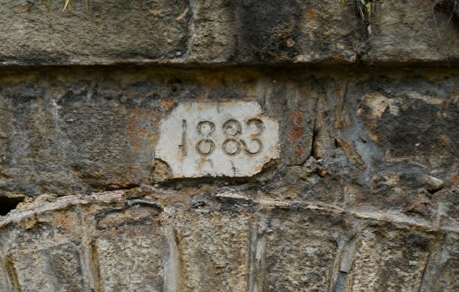 Fotografia em close de uma placa de pedra clara encrustada em uma parede de rocha bruta escura. Na placa estão esculpidos os numerais mil oitocentos e oitenta e três indicando o ano de mil oitocentos e oitenta e três. A pedra ao redor apresenta texturas irregulares e sinais de desgaste natural pelo tempo.