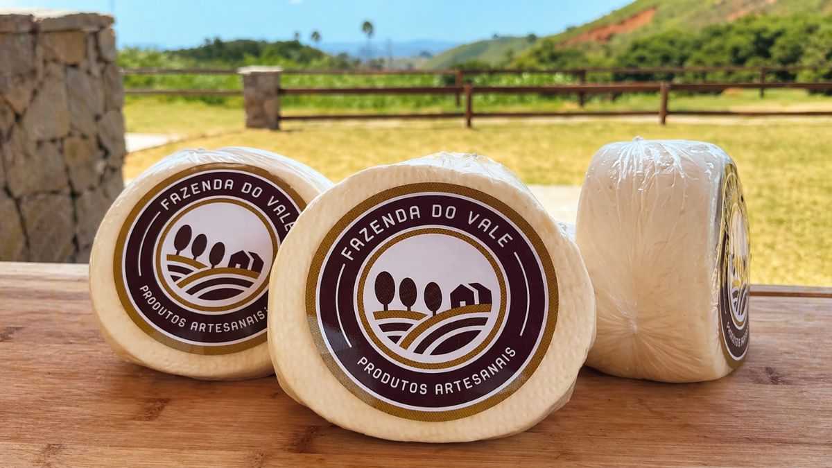 Uma fotografia em primeiro plano mostrando três peças de queijo artesanal redondas sobre uma tábua de madeira clara. Os queijos estão embalados em plástico filme transparente e possuem um rótulo circular central com a inscrição Fazenda do Vale Produtos Artesanais em tons de marrom e dourado. Ao fundo desfocado, observa-se uma paisagem rural com gramado verde, cercas de madeira e montanhas sob um céu azul claro, destacando a origem campestre do produto.