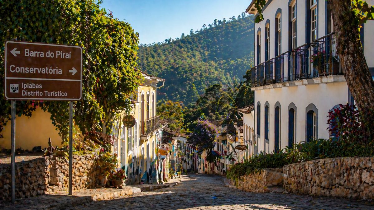 Uma vista de uma rua estreita e íngreme de paralelepípedos na cidade de Ipiabas, Barra do Piraí, Rio de Janeiro. À esquerda, uma placa de sinalização de trânsito marrom com letras brancas indica as direções para Barra do Piraí (seta para a esquerda) e Conservatória (seta para a direita), com o texto abaixo: Ipiabas Distrito Turístico. A rua é ladeada por edifícios coloniais de dois andares, com fachadas caiadas de branco e janelas com molduras coloridas, muitas com varandas de ferro forjado. As construções descem a colina em direção a colinas verdes e exuberantes cobertas por floresta densa. O céu é azul e claro, com luz solar brilhante iluminando a cena. A rua está vazia de pessoas ou veículos. Muros de pedra rústica revestem a calçada no primeiro plano.