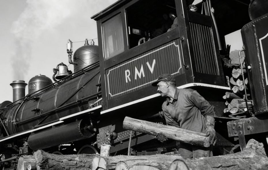 Fotografia em preto e branco de um operário ferroviário de perfil segurando uma grande tora de madeira para abastecer uma locomotiva a vapor. Na lateral da cabine do trem aparecem as letras iniciais RMV em branco sobre fundo escuro. Há pilhas de lenha em primeiro plano e o céu está claro com um pouco de fumaça saindo da chaminé da máquina.