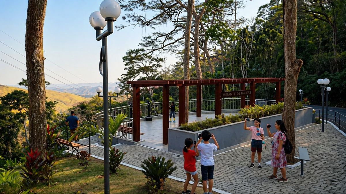 Uma fotografia colorida do Mirante de Ipiabas durante o dia. No primeiro plano, à direita, quatro crianças e jovens estão em pé sobre um caminho de bloquetes cinzas segurando celulares para fotografar a paisagem. À esquerda, um poste de iluminação preto com dois globos brancos redondos se destaca. No centro da imagem, há uma estrutura de pergolado em madeira avermelhada sobre um deck de piso claro, cercado por guarda-corpos metálicos. Uma placa no pergolado identifica o local como Mirante de Ipiabas. Ao fundo, um homem observa o horizonte sentado em um banco de madeira. A paisagem ao redor é composta por montanhas onduladas sob um céu azul claro e vegetação densa de Mata Atlântica com árvores altas que emolduram a cena. A luz do sol é suave, sugerindo o período da tarde.