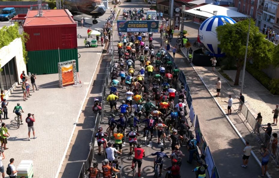 Vista aérea da largada do Bike Fest em Ipiabas, com centenas de ciclistas preenchendo a rua. Ao fundo, um avião decorativo e um balão inflável compõem o cenário do evento, que é acompanhado por espectadores nas laterais.