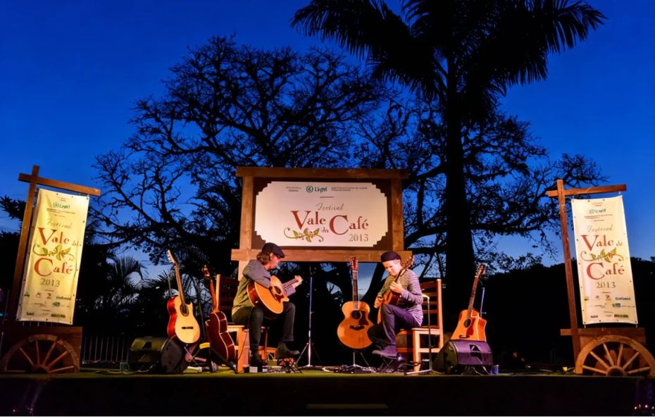 Dois músicos sentados em um palco de madeira ao ar livre tocando violões acústicos durante o Festival Vale do Café. Ao fundo há uma grande árvore e o céu azul do entardecer. No palco há banners e uma placa central com o logotipo oficial do evento.