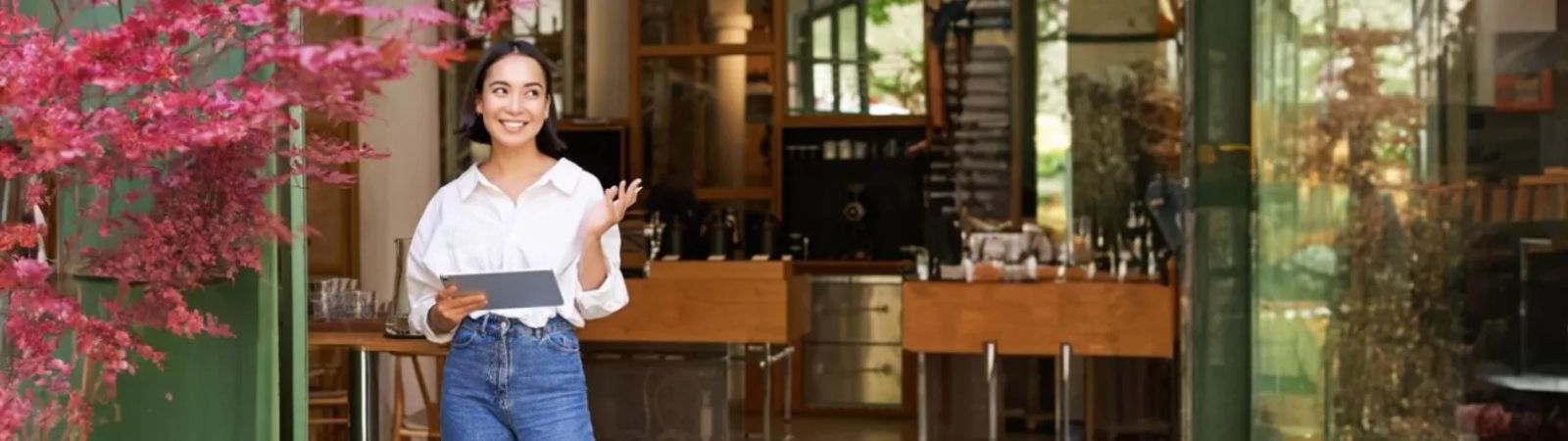 Uma mulher sorridente em frente a um restaurante moderno com decoração em madeira e uma planta de folhas avermelhadas na entrada.