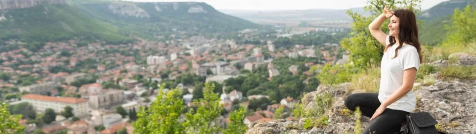 Mulher sentada sobre uma grande pedra no topo de um morro observando a vista panorâmica do distrito de Ipiabas com suas casas e montanhas ao fundo sob um céu claro.