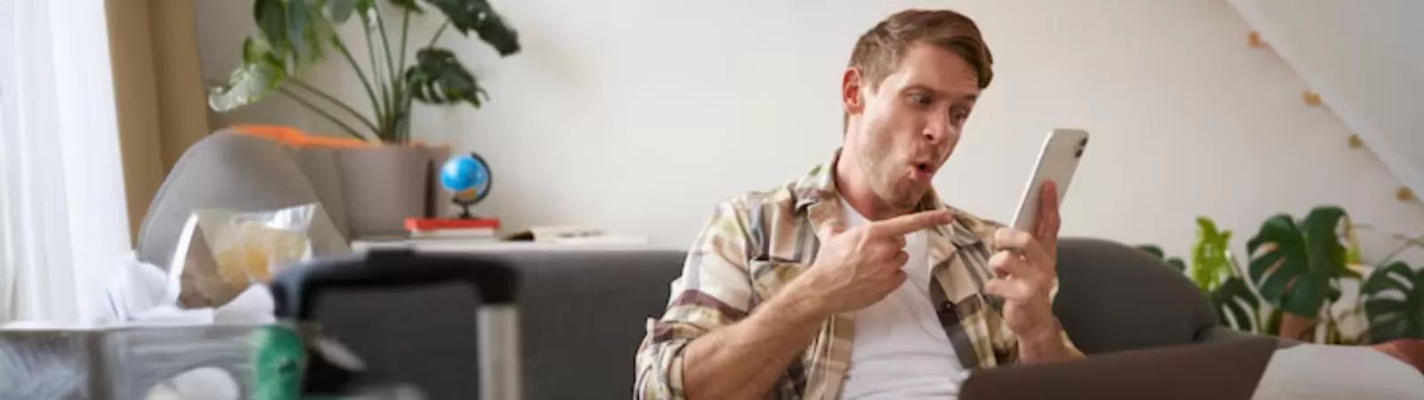 Um homem jovem com expressão de surpresa e alegria está sentado em um sofá cinza segurando um smartphone branco. Ele aponta para a tela do aparelho com o dedo indicador da mão direita. Ao fundo aparece uma sala iluminada com plantas verdes e malas de viagem indicando que ele está planejando um passeio.