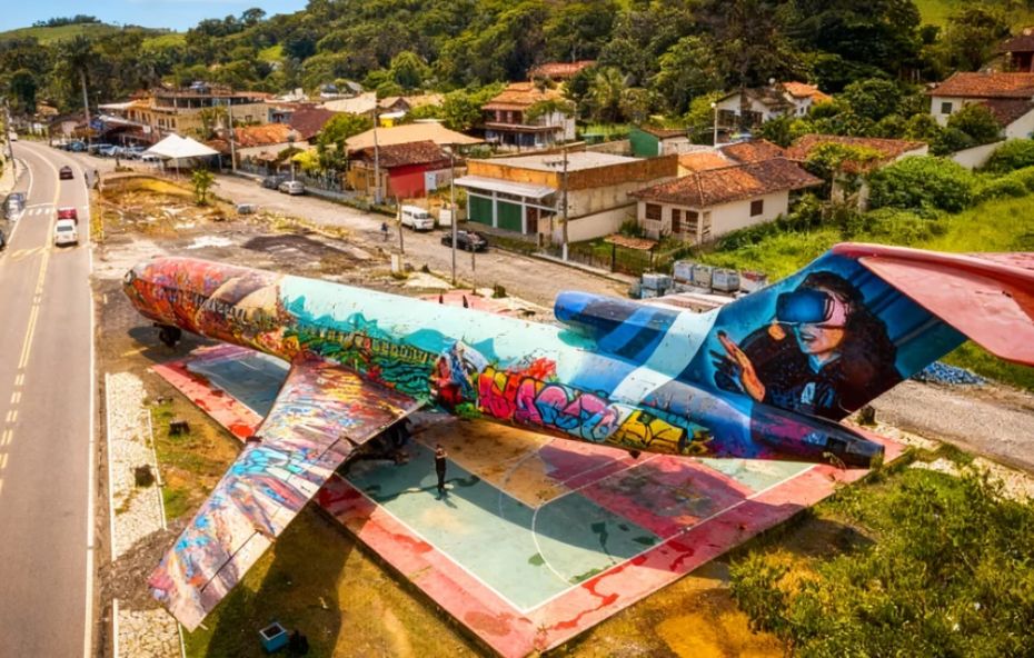 Vista aérea de um grande avião estacionado em Ipiabas, totalmente coberto por arte em grafite. Ao fundo, veem-se a estrada e as casas do distrito, cercadas por vegetação.