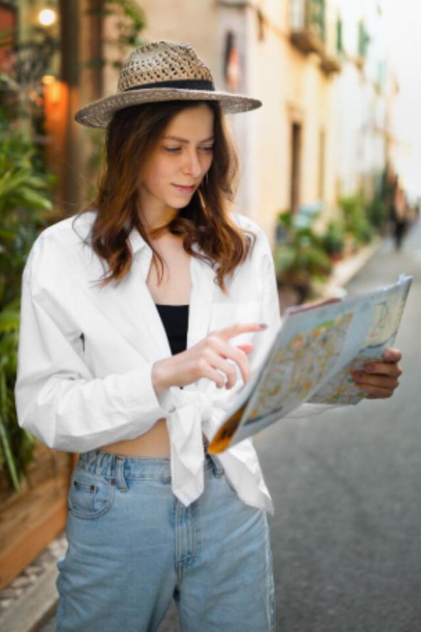 Jovem turista vestindo chapéu de palha e camisa branca aberta sobre blusa preta segurando um mapa aberto em uma rua charmosa e arborizada.
