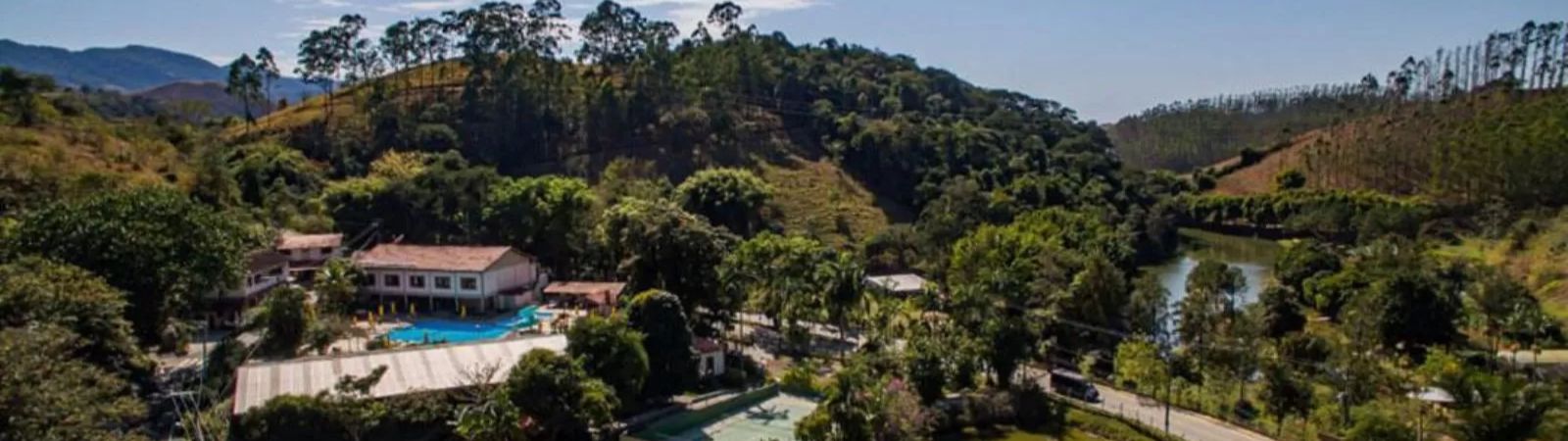 Vista aérea de um hotel fazenda em Ipiabas cercado por montanhas e áreas verdes com piscina quadra de tênis e um lago sereno refletindo o céu.