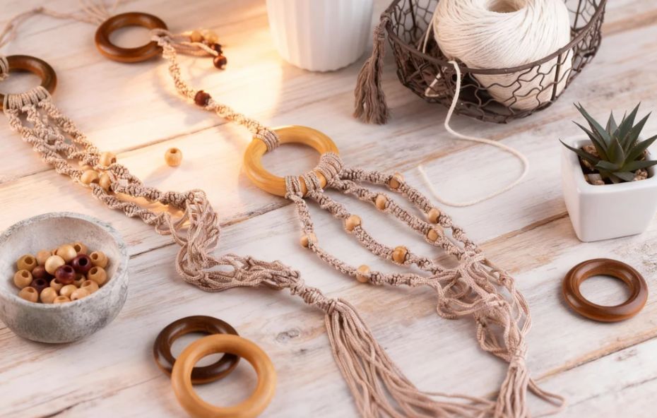 Uma mesa de madeira clara com luz solar suave onde repousam peças de macramê em processo de produção. A imagem mostra fios trançados em tons de bege, argolas de madeira clara e escura, contas de madeira em um pequeno recipiente de cerâmica, uma cesta de metal com um novelo de linha branca e um pequeno vaso com uma planta suculenta.