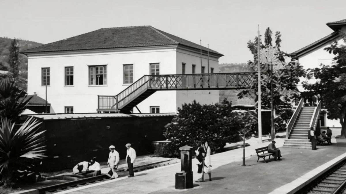 Fotografia antiga em preto e branco mostrando uma passarela metálica elevada conectando prédios ferroviários com pessoas na plataforma de embarque.