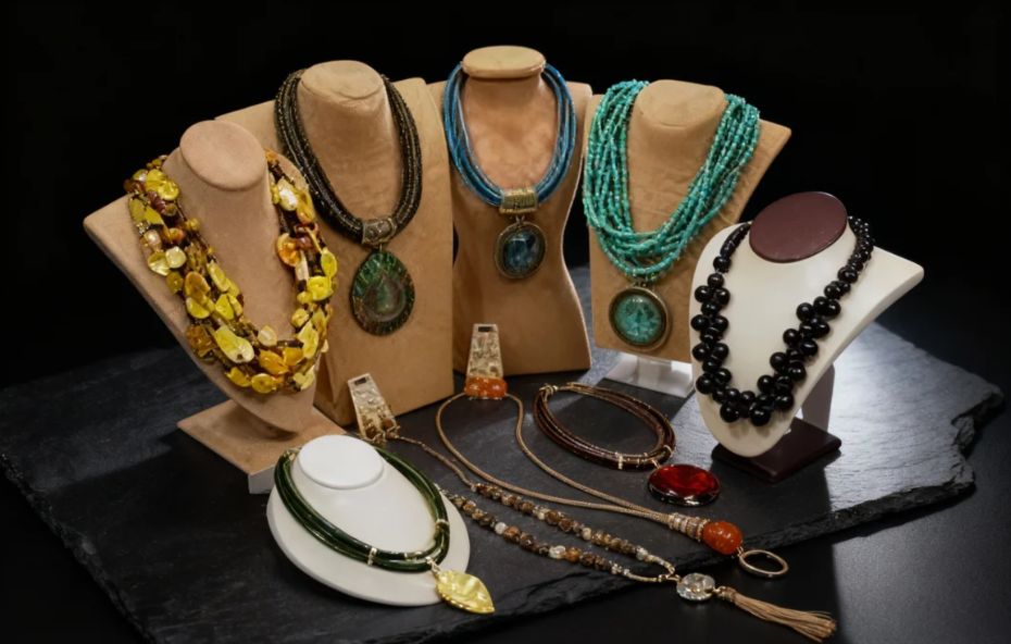 Uma coleção de colares artesanais de luxo expostos em bustos de camurça e suportes brancos sobre uma base de pedra escura. As peças apresentam diversos materiais como pedras naturais em tons de turquesa e âmbar, pingentes circulares grandes, contas pretas brilhantes, elementos metálicos e detalhes em cordão. A iluminação destaca as texturas e as cores vibrantes dos acessórios.
