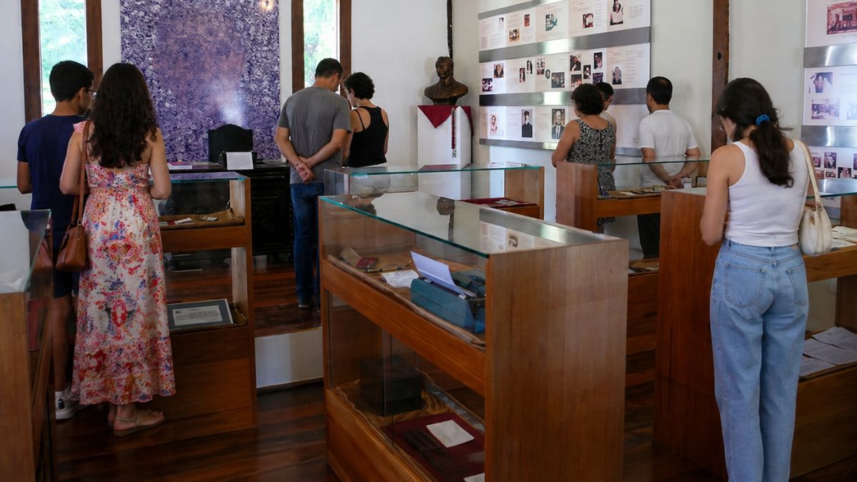 Visitantes de costas observam fotos e documentos em painéis e vitrines de madeira no interior do Memorial Aracy Carvalho Di Biase.