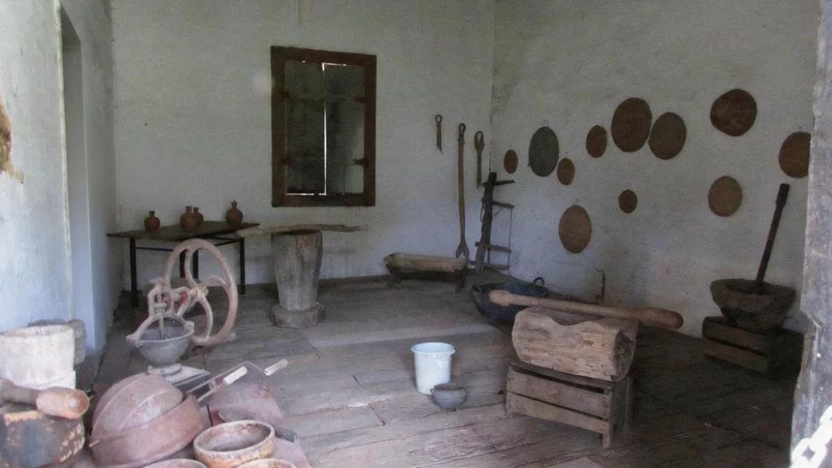 Sala histórica com paredes brancas exibindo pilões de madeira, peneiras de palha na parede e diversos utensílios domésticos rústicos.