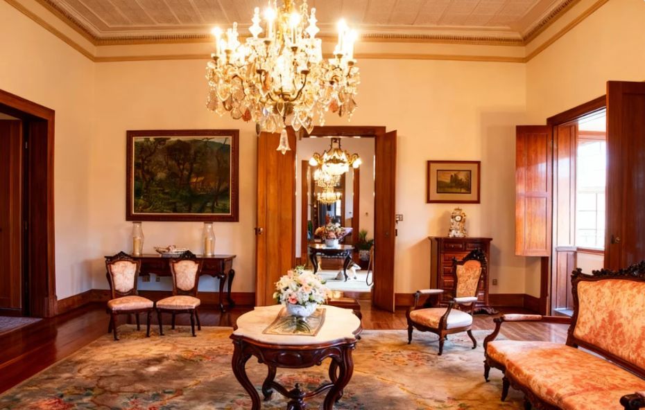 Visão de um salão imperial com sofás de madeira entalhada e estofamento rosa decorado com flores além de poltronas vermelhas e uma mesa circular de tampo de mármore posicionada sob um lustre de cristal com velas acesas em primeiro plano.