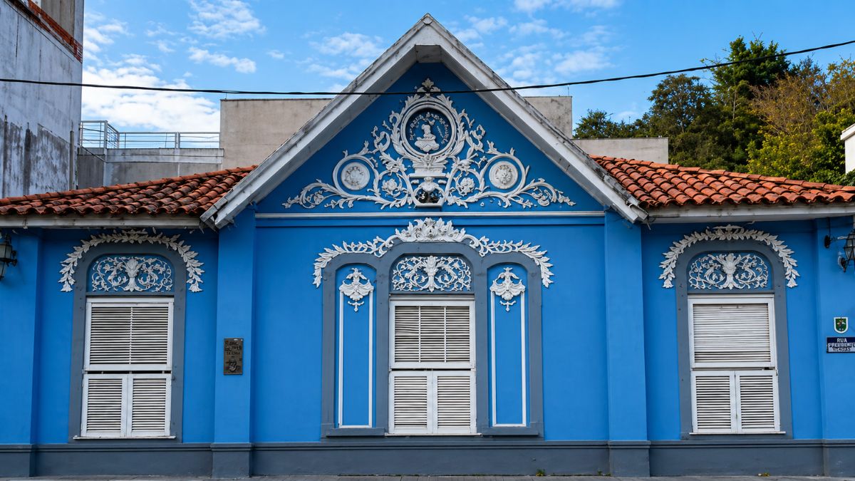 Fotografia frontal de uma fachada histórica em azul vibrante com ornamentos brancos em alto relevo. O destaque central é um frontão triangular que exibe o medalhão da Princesa Isabel e elementos heráldicos. Três janelas com venezianas brancas e molduras decoradas por guirlandas e volutas compõem a simetria da edificação sob um telhado de telhas cerâmicas e céu aberto.