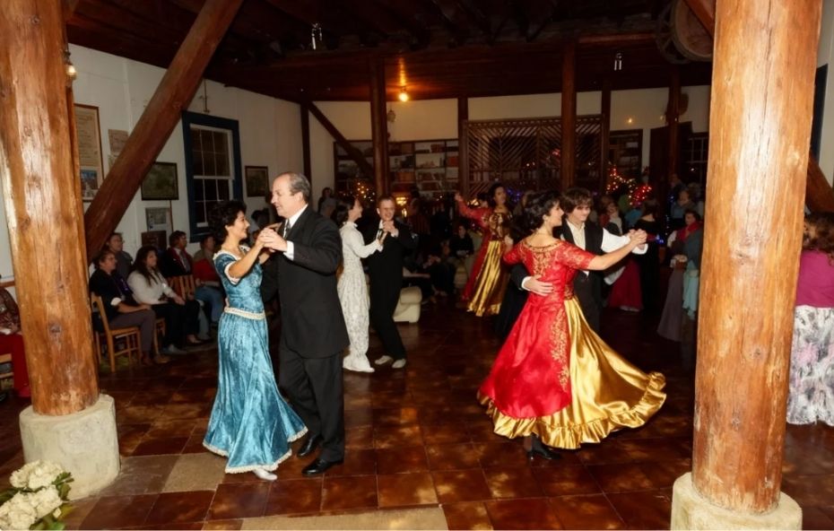 Fotografia de casais dançando em um salão amplo com pilares de madeira, vestindo trajes clássicos do século dezenove, com mulheres em vestidos longos e rodados em tons de azul e vermelho com dourado, enquanto pessoas assistem ao fundo.