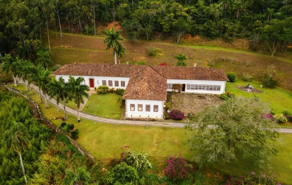 Fotografia de um salão interno com teto de madeira e paredes brancas decoradas com quadros de época e um grande lustre de cristal centralizado sobre móveis clássicos em tons de rosa e vermelho dispostos em um tapete estampado sob a luz de velas.