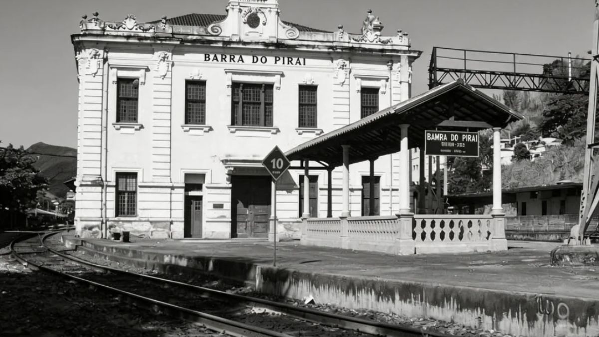 Fotografia em preto e branco da fachada lateral da Estação Ferroviária de Barra do Piraí mostrando trilhos e plataforma de embarque.