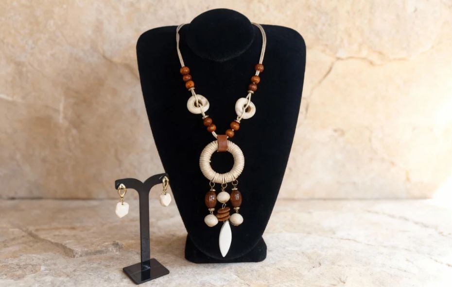 Fotografia em plano médio de um conjunto artesanal composto por um colar robusto e brincos. O colar apresenta um pendente circular revestido em palha clara com esferas de madeira marrom e detalhes em marfim vegetal sobre um busto de veludo preto. Os brincos pequenos em formato de gota clara estão em um suporte lateral.