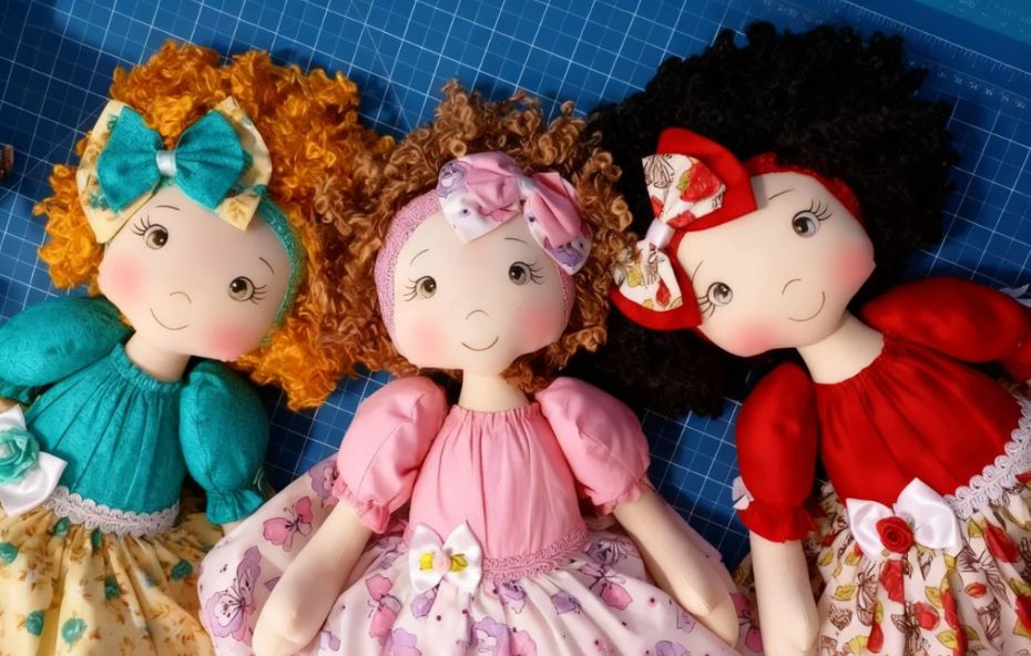 Produção artesanal de três bonecas de pano com cabelos cacheados volumosos e vestidos coloridos representando a tradição artística de Ipiabas sobre uma base de corte técnica.