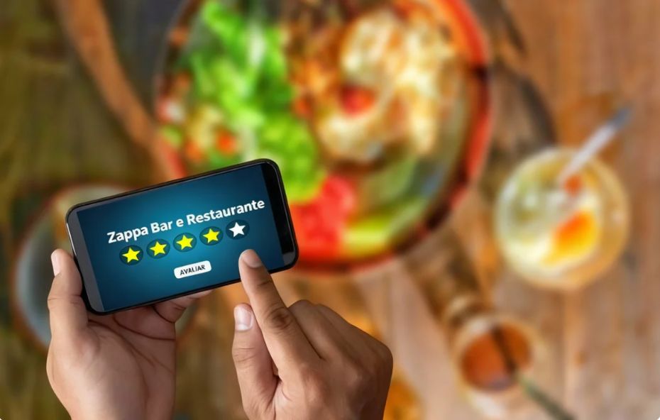 Mão segurando um smartphone que exibe quatro estrelas amarelas e uma branca para o Zappa Bar e Restaurante sobre um fundo desfocado de comida e bebidas.