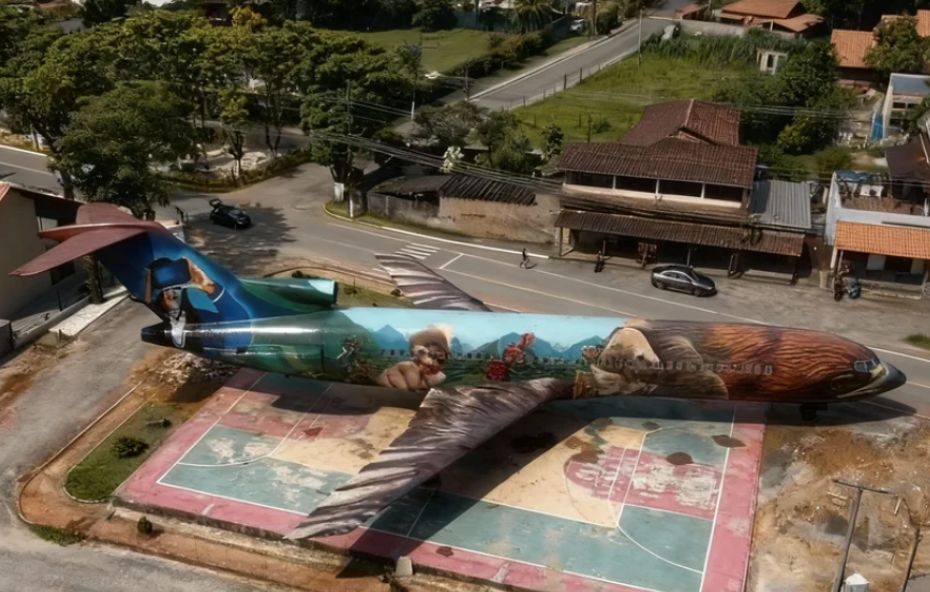 Vista aérea de um avião de grande porte estacionado em uma praça colorida no Centro de Ipiabas. A aeronave possui uma pintura artística detalhada em toda a sua fuselagem com figuras que remetem à cultura local e está posicionada ao lado de uma via urbana com casas ao fundo.
