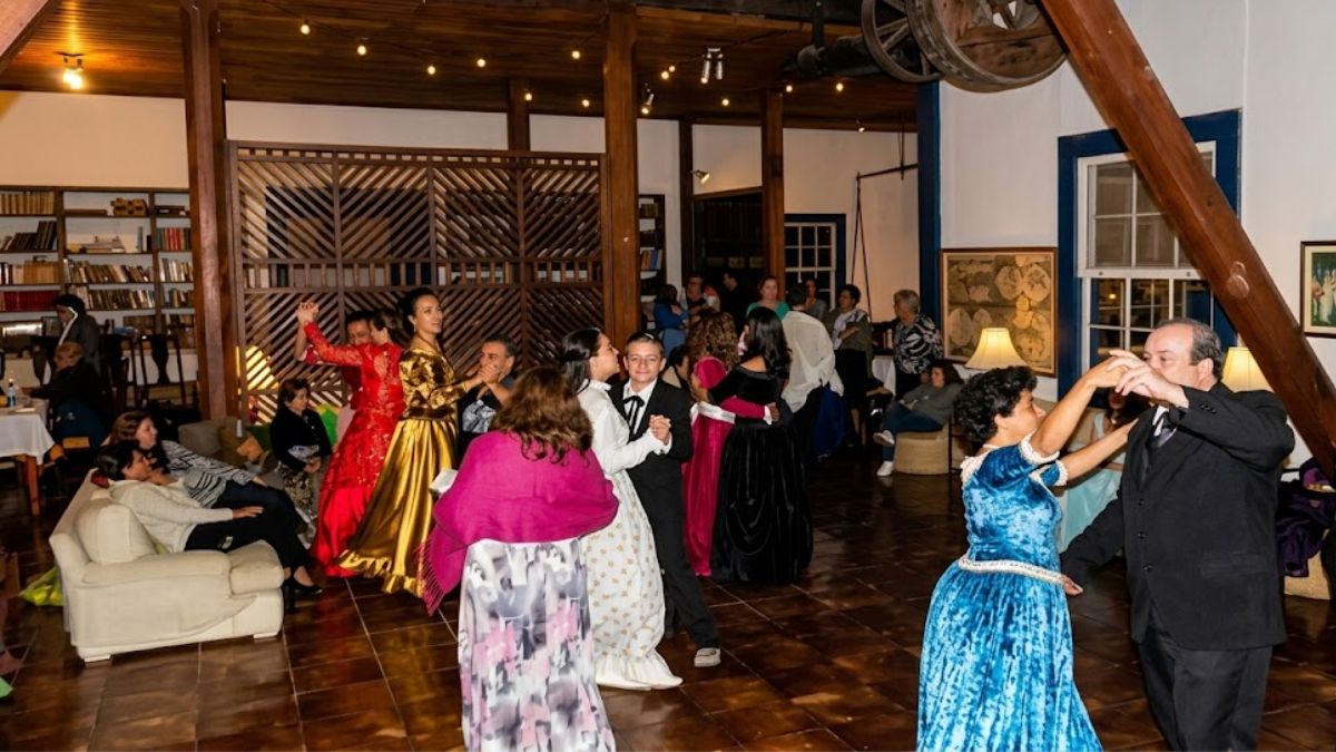 Pessoas dançando em um salão rústico com trajes de gala antigos, vestidos coloridos e ternos, sob luzes quentes e teto de madeira.