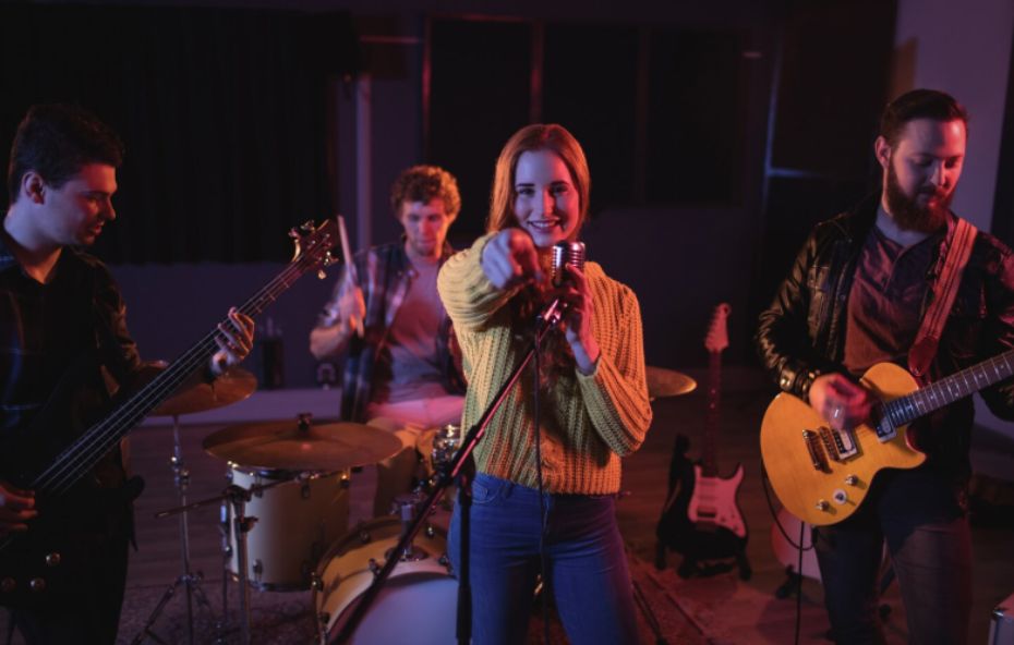 Uma banda de quatro integrantes se apresenta em um palco com iluminação roxa e azul, destacando uma vocalista sorridente ao centro, um guitarrista, um baixista e um baterista ao fundo.