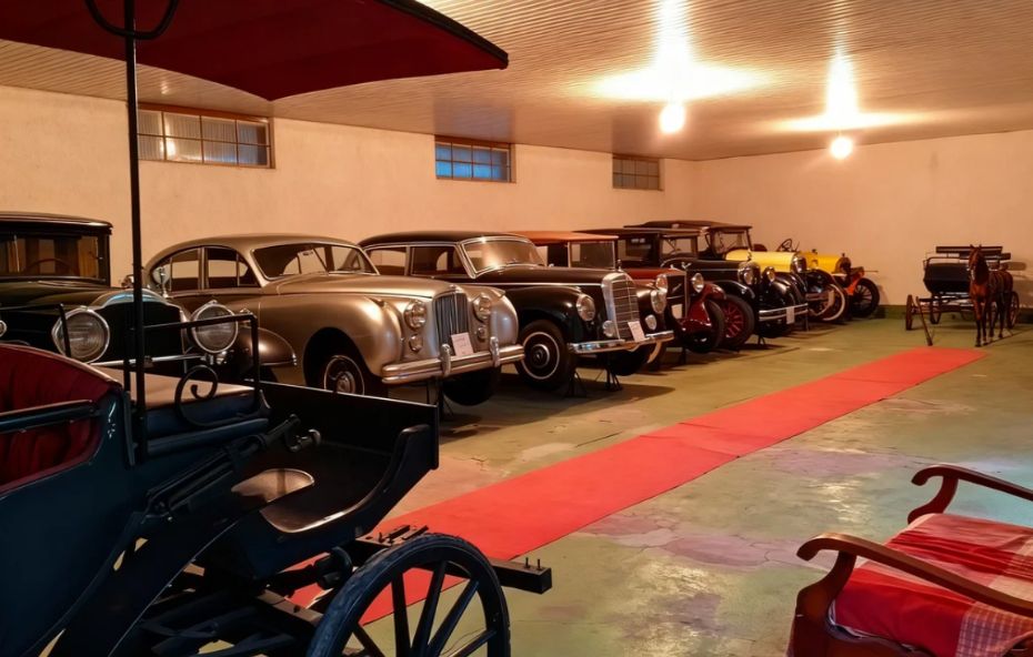 Fotografia de um galpão amplo exibindo uma fileira de carros antigos de luxo das décadas de quarenta e cinquenta com destaque para modelos pretos e prateados estacionados sobre um piso verde ao lado de uma passarela vermelha e uma carruagem histórica.