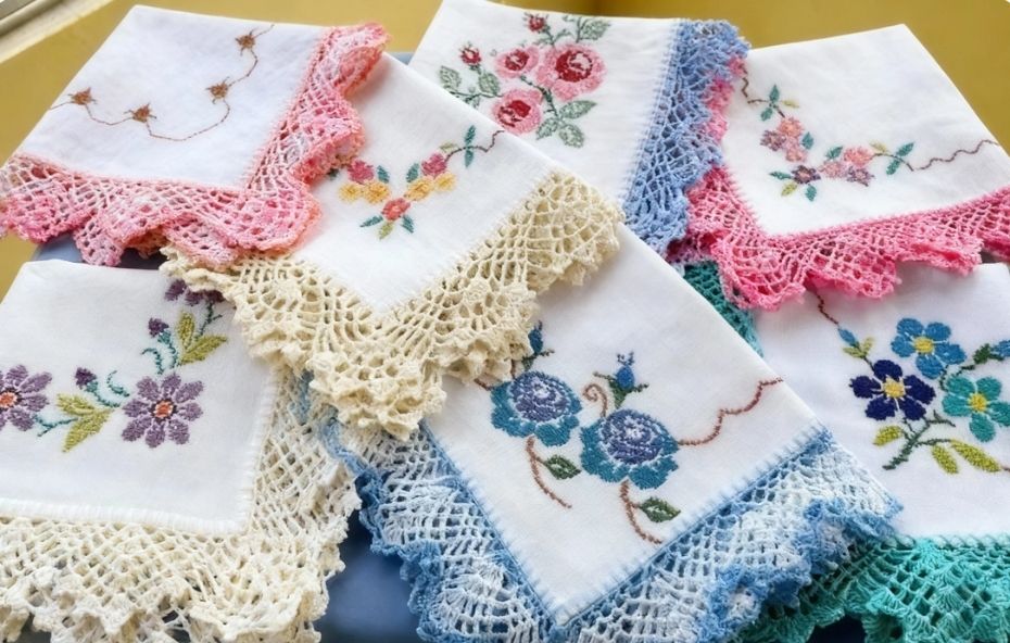 Vários lenços de tecido branco dobrados exibindo flores bordadas em ponto cruz nas cores azul, rosa e lilás com acabamento em bicos de crochê coloridos.